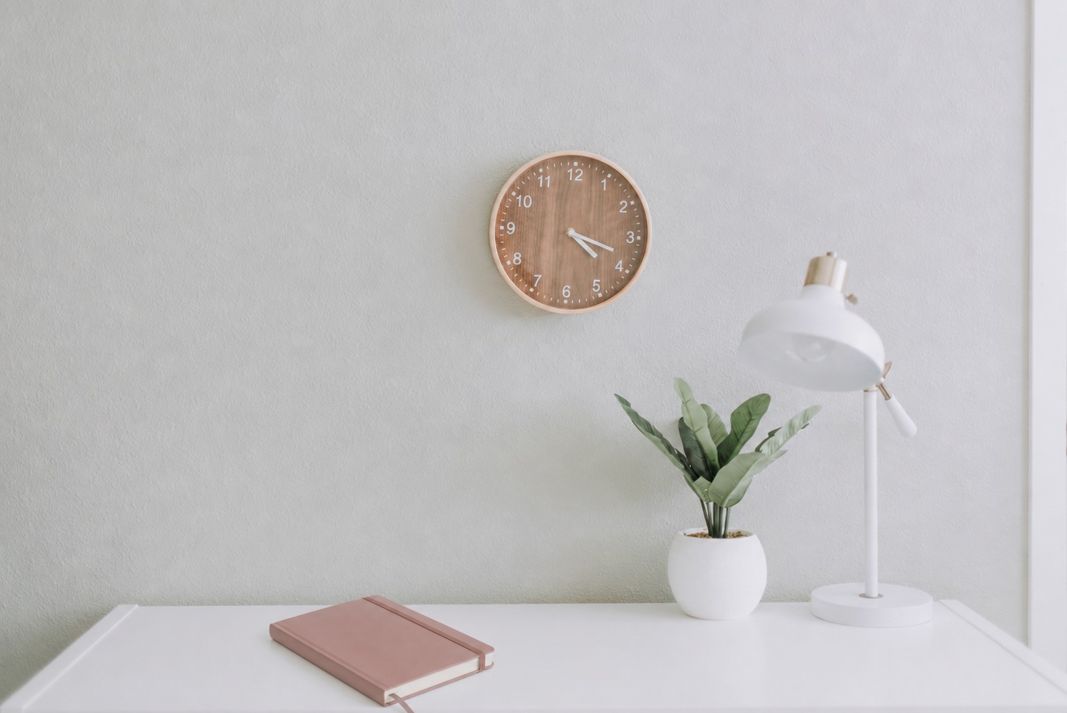 Heller Schreibtisch mit Wanduhr aus Holz, Tischlampe, Pflanze und rosa Notizbuch